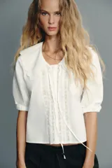 Camisa blanca de cuello bobo con bordados, escote pico con lazada, manga corta abullonada con elástico y cierre frontal con botones.