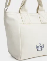 Cartera mini de lona color crudo con estampado "The Brunch Club" en azul marino. Tiene correa regulable y desmontable, manijas superiores y cierre superior.