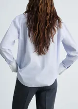 Camisa celeste a rayas verticales, de corte recto y manga larga con puños abotonados en blanco. Presenta cuello camisero blanco, cierre frontal con botones y un bolsillo de parche en el pecho.