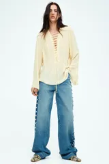 Blusa color crema de gasa con escote en V y cordones cruzados. Tiene mangas largas acampanadas y pliegues en la cintura.