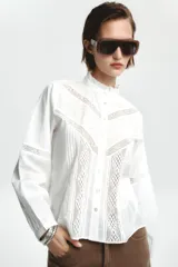 Camisa blanca de cuello alto y manga larga, con detalle de entredós combinados a tono y cierre frontal con botones.