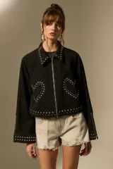 Campera cropped de jean color negro, con cuello camisero, cierre frontal y detalles de tachas plateadas en el cuello, hombros, mangas y ruedo. Presenta un diseño de tachas en forma de corazón en el pecho.