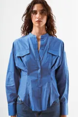 Camisa de manga larga color azul con finas rayas verticales blancas. Presenta cuello mao, botones frontales y dos bolsillos de parche con tapa en el pecho. La prenda tiene un diseño ajustado en la cintura con pinzas y un volumen holgado en las mangas.