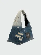Bolso de hombro Marc Jacobs modelo The Sack Bag, confeccionado en denim azul con efecto desgastado y roturas. Presenta el logo de la marca estampado en el frente.