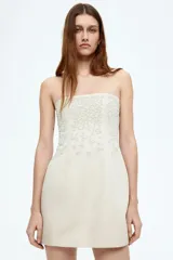 Vestido corto strapless color beige, confeccionado en mezcla de lino y viscosa, con bordado de nudo francés tono sobre tono en la parte superior.