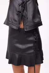 Falda corta negra de efecto cuero, estilo wrap o cruzada, con ajuste lateral mediante lazos de cuero y terminación con volante o ruedo con volado. Presenta costuras contrastantes en color claro.