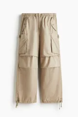 Pantalón cargo de algodón color beige, de corte holgado y tiro medio. Presenta cintura elástica con cordón ajustable, bolsillos laterales y grandes bolsillos cargo con solapa en las piernas. Los bajos son ajustables con cordón oculto.