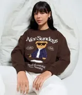 Buzo de algodón y felpa color marrón oscuro con cuello redondo y mangas largas. Presenta un estampado frontal de un oso con gafas de sol y traje a rayas, con el texto "Nice Sundays" en la parte superior y la frase "un café si vous plait morning" en la parte inferior.