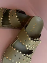 Sandalia tipo mule de mujer, color dorado metalizado, con dos tiras anchas superpuestas con borde festoneado y tachas doradas. La suela es gruesa y de color marrón claro.