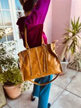 Bolso de mano grande de cuero color marrón, con doble asa corta y correa larga desmontable. Tiene cierre superior y detalles de flecos en el frente.