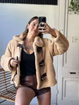 Campera corta color beige de tela tipo peluche con cuello solapa y cierre frontal con presillas de cuero sintético y hebillas metálicas.