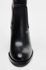 Bota alta de cuero sintético negro con caña alta y tacón ancho. Detalle de hebilla en la parte superior de la caña.