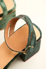 Sandalia de terciopelo verde con taco cuadrado de 7 cm, punta cuadrada y tiras anchas anudadas en el empeine. Cuenta con una tira fina ajustable al tobillo con hebilla dorada.