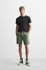 Bermuda color verde militar de corte relajado, confeccionada en tejido de algodón. Tiene cintura elástica ajustable con cordón interior, bolsillos frontales y bolsillo trasero de parche.
