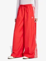 Pantalón Adidas Firebird adicolor oversized de color rojo con las tres tiras blancas a los costados. Cuenta con bolsillos con cremallera y cintura con cordón ajustable.