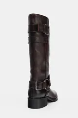 Bota de caña alta de cuero color marrón oscuro, estilo motociclista, con taco bajo y punta redonda. Presenta una tira con hebilla ajustable en la parte superior de la caña y una tira desmontable con argolla metálica y detalle de rosa repujada en el tobillo.