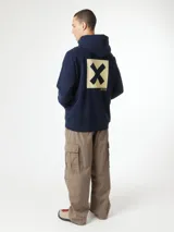 Buzo hoodie azul marino con capucha y cordones ajustables, de corte holgado y puños elásticos. Presenta un estampado en la espalda con una cruz dorada dentro de un cuadrado y texto en japonés.