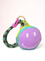 Llavero monedero redondo con diseño de cara sonriente en color lila y celeste. Tiene cierre superior con cremallera amarilla, mosquetón de cuerda multicolor y charms colgantes.