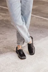 Mocasín de cuero negro con cordones y tachas metálicas en la puntera.