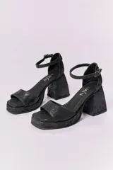 Sandalias de cuero negro con plataforma y tacón cuadrado alto. Presentan una tira delgada sobre los dedos y pulsera tobillera con hebilla dorada.