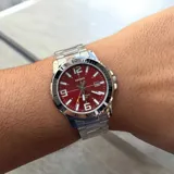 Reloj análogo Casio con caja y pulsera de acero inoxidable plateado. Presenta una esfera redonda color rojo vibrante con marcadores de hora grandes y luminosos, números arábigos en 12, 9, 6 y 3, y ventana de fecha a las 3. El bisel plateado tiene marcadores de minutos. Posee tres manecillas plateadas y una manecilla de segundos con punta amarilla.