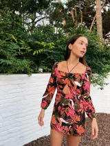 Minivestido marrón con estampado floral en tonos naranja, rosa y verde. Tiene escote cuadrado con abertura en el centro, mangas largas y falda con tajo lateral.