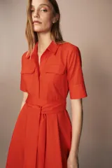 Vestido midi camisero color ladrillo, confeccionado en popelina de algodón. Presenta cuello solapa, manga corta con puños abotonados, bolsillos laterales ocultos y bolsillos de parche con solapa en el pecho. Incluye cinturón lazada en el mismo tejido y cierre frontal con botones ocultos.