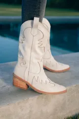 Bota texana de caña alta color marrón claro, confeccionada en cuero vacuno con detalles de bordado y apliques en relieve. Tiene cierre lateral, taco de madera con folia de cuero y suela de goma. La horma es de punta fina, ancha y cómoda.