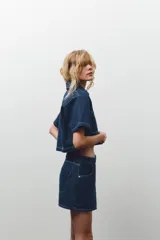 Camisa corta de denim azul con cuello solapa, escote pico, manga corta, bolsillos delanteros de plastrón y cierre frontal con botones metálicos.