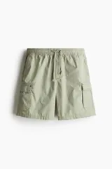 Short cargo color verde claro en popelina de algodón, con cintura elástica ajustable con cordón, bolsillos laterales, bolsillos cargo con solapa y botón a presión en piernas y traseros, y aberturas laterales en el bajo.