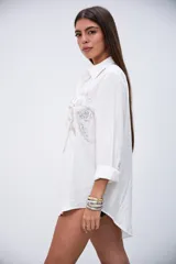 Camisa oversize color crudo con cuello clásico, cierre de botones frontales y mangas largas con puños. Presenta bordados florales con lentejuelas y mostacillas plateadas en la parte delantera.