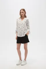 Blusa color beige con estampado de arabescos blancos, cuello mao, mangas 3/4 con lazos en los puños, cierre parcial con botones y bolsillos frontales.