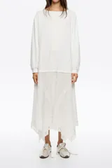 Vestido largo blanco de manga larga y escote barco. La parte superior es de tejido liso y la falda es fluida, con detalles de encaje en forma de V y bajo asimétrico con terminación de encaje.