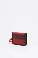 Billetera compacta de color rojo con ribetes en contraste de color marrón oscuro. Presenta el logo de Bimba y Lola estampado en blanco en el frente y una correa de muñeca desmontable sujeta con un mosquetón metálico.