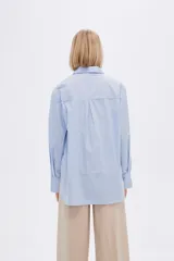 Camisa a rayas verticales verdes y blancas, de corte holgado, manga larga con puños abotonados, cuello clásico y bolsillo de parche en el pecho.