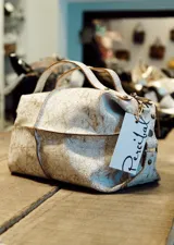 Bolso de cuero con efecto craquelado en tonos blanco y beige, de diseño amplio con doble asa superior y cierre de cremallera extendido. Presenta detalles de tachas metálicas en los laterales y un estilo de inspiración bohemia.