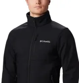 Chaqueta softshell Columbia para hombre, color negro, con cierre frontal, cuello alto y logo de la marca en blanco en el pecho. Presenta bolsillos laterales y uno en el pecho con cierre.