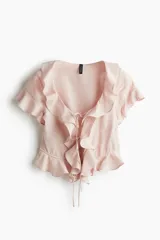Blusa color rosa pálido de lino y viscosa, con escote en V pronunciado, mangas cortas y volados en el escote, mangas y ruedo. Se ata en el centro con dos cordones finos.