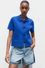 Chaqueta de punto azul con cuello solapa y manga corta. Tiene bolsillos delanteros de parche y cierre frontal con botones grandes con abalorios.