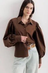Camisa marrón con cuello clásico, abotonada al frente y mangas largas abullonadas con puños abotonados. Corte recto.