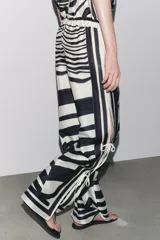 Pantalón de tiro medio con cintura elástica y pierna ancha. Presenta un estampado de líneas onduladas en blanco y negro, y aberturas laterales ajustables con lazadas.
