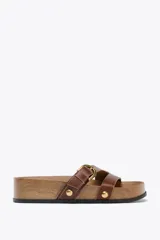 Sandalia plana de cuero color marrón con plataforma de madera. Tiene tiras anchas en la parte delantera con detalles metálicos y cierre mediante hebilla. La suela tiene efecto acolchado y la punta es redonda.