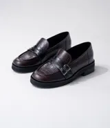 Mocasines de cuero color marrón con estampado de cocodrilo, detalle de hebilla metálica plateada y suela de goma negra. Diseño clásico con punta redonda y calce cómodo.