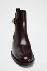 Botín chelsea color marrón oscuro de cuero sintético con elásticos laterales, hebilla decorativa dorada y tirador trasero.