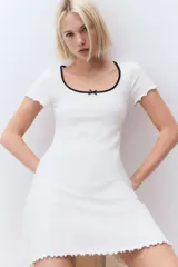 Vestido mini blanco de manga corta en punto de algodón acanalado con pollera evasé. Modelo con cuerpo entallado y escote redondo amplio con ribete fino en color negro y moña decorativa de satén. Ribete sobrehilado en mangas y bajo.