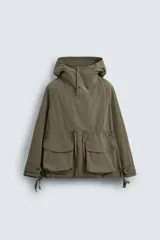 Parka color verde militar de corte amplio, confeccionada en tejido técnico ligero. Cuenta con cuello alto con capucha ajustable, cierre frontal con botones, mangas largas con puños ajustables, bolsillos de plastrón con solapa en la parte delantera, cintura ajustable con cordón y bajo con aberturas laterales ajustables con cordón.
