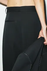 Pantalón negro de tiro medio con trabillas, falso bolsillo de vivo en espalda, detalle de pespuntes combinados a contraste y pinzas. Cierre frontal con cremallera y botón.