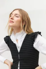 Chaleco blanco con finas rayas verticales negras, escote en V, cierre frontal con botones negros y volados en lino negro en los hombros. La parte inferior presenta otro volado en la misma tela del cuerpo.