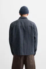 Camisa de hombre a cuadros azules y negros, de corte cuadrado, con cuello solapa, manga larga con puños abotonados, cierre frontal con botones y bolsillos de parche con solapa en el pecho.