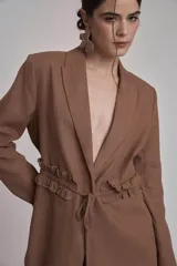Blazer marrón con solapas de muesca, cierre frontal con lazo y mangas largas. Presenta volados a lo largo de las costuras laterales del pantalón a juego.